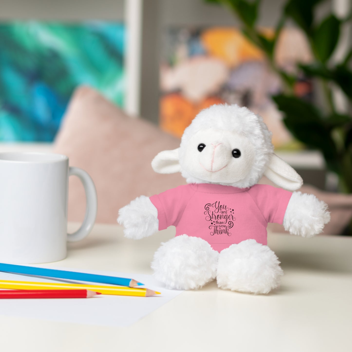 White plush lamb toy wearing a pink shirt with text, sitting on a table with a mug and pencils.