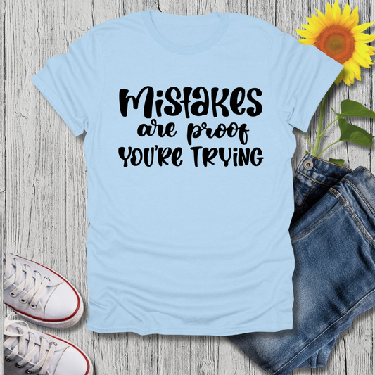 Light blue t-shirt with 'Mistakes are proof you're trying' text, sunflower, jeans, and sneakers on a wooden surface.