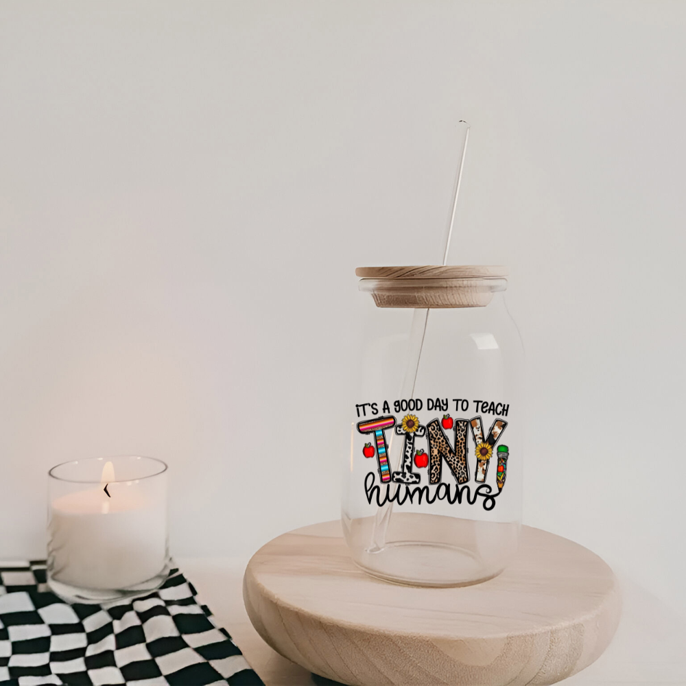 Glass tumbler with straw lid on a wooden surface next to a candle, against a white background.