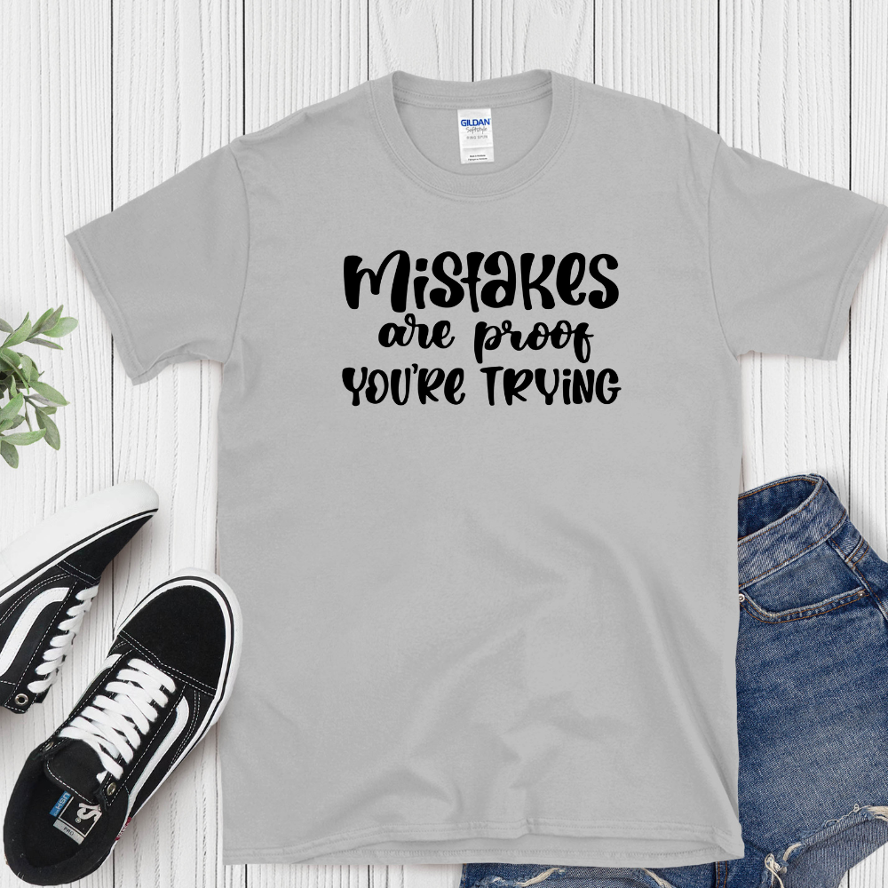 Gray t-shirt with 'Mistakes are proof you're trying' text, paired with sneakers and jeans on a wooden surface.