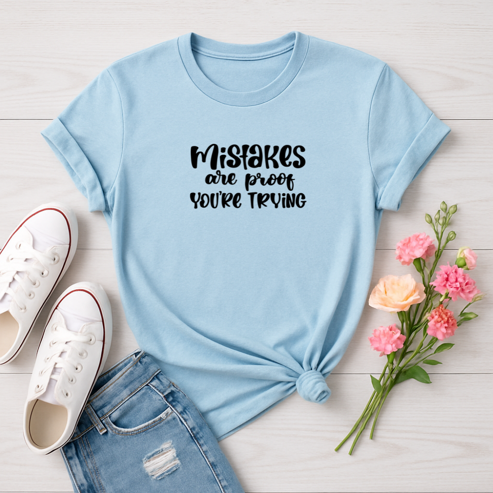 Light blue t-shirt with 'Mistakes are proof you're trying' text, paired with white sneakers and pink flowers on a light wooden surface.