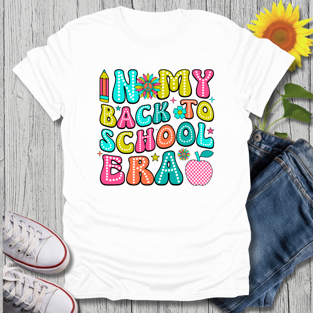 White t-shirt with colorful 'In My Back to School Era' text, paired with jeans and sneakers on a wooden surface.