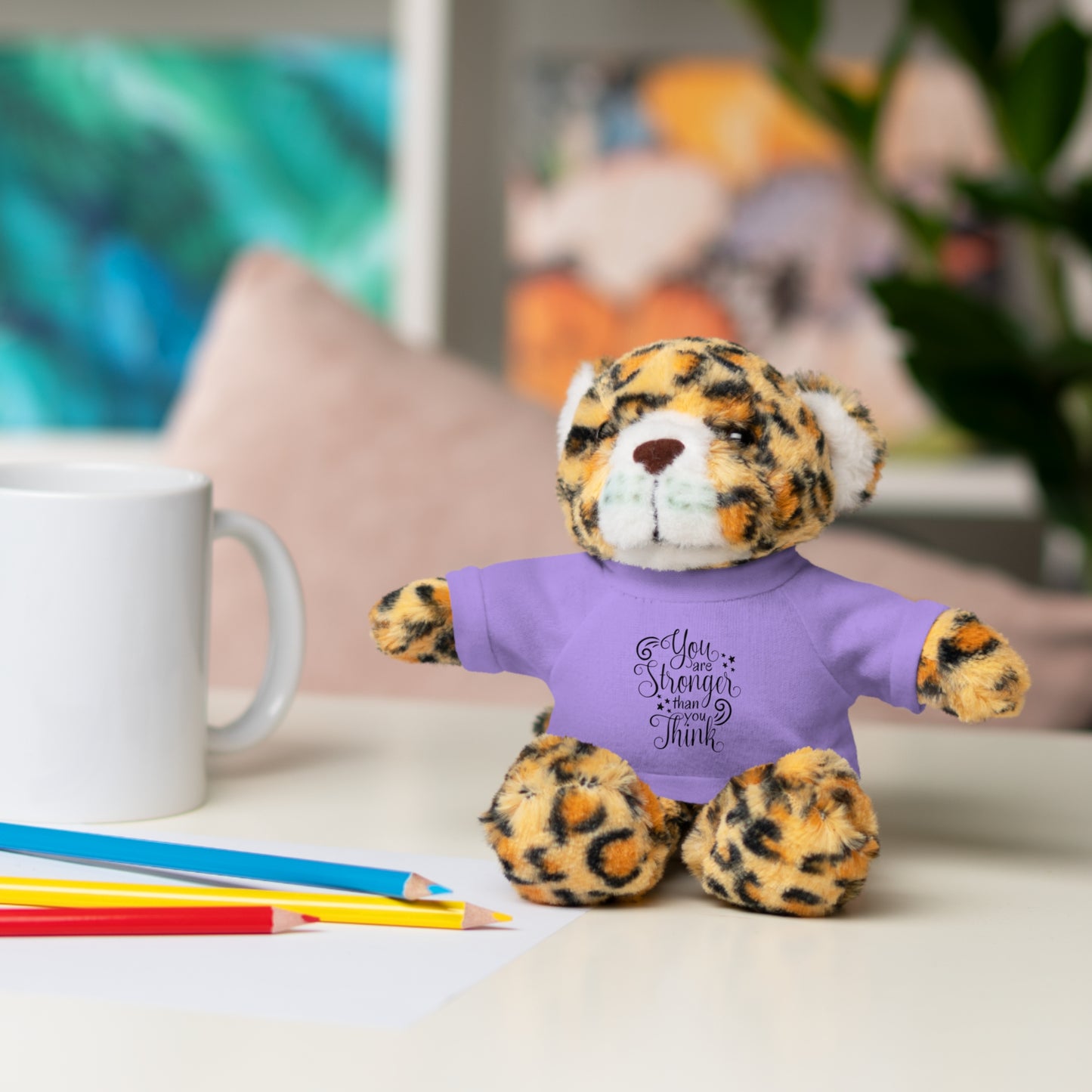 Leopard print teddy bear wearing a purple shirt with text, sitting on a desk with a mug and pencils.