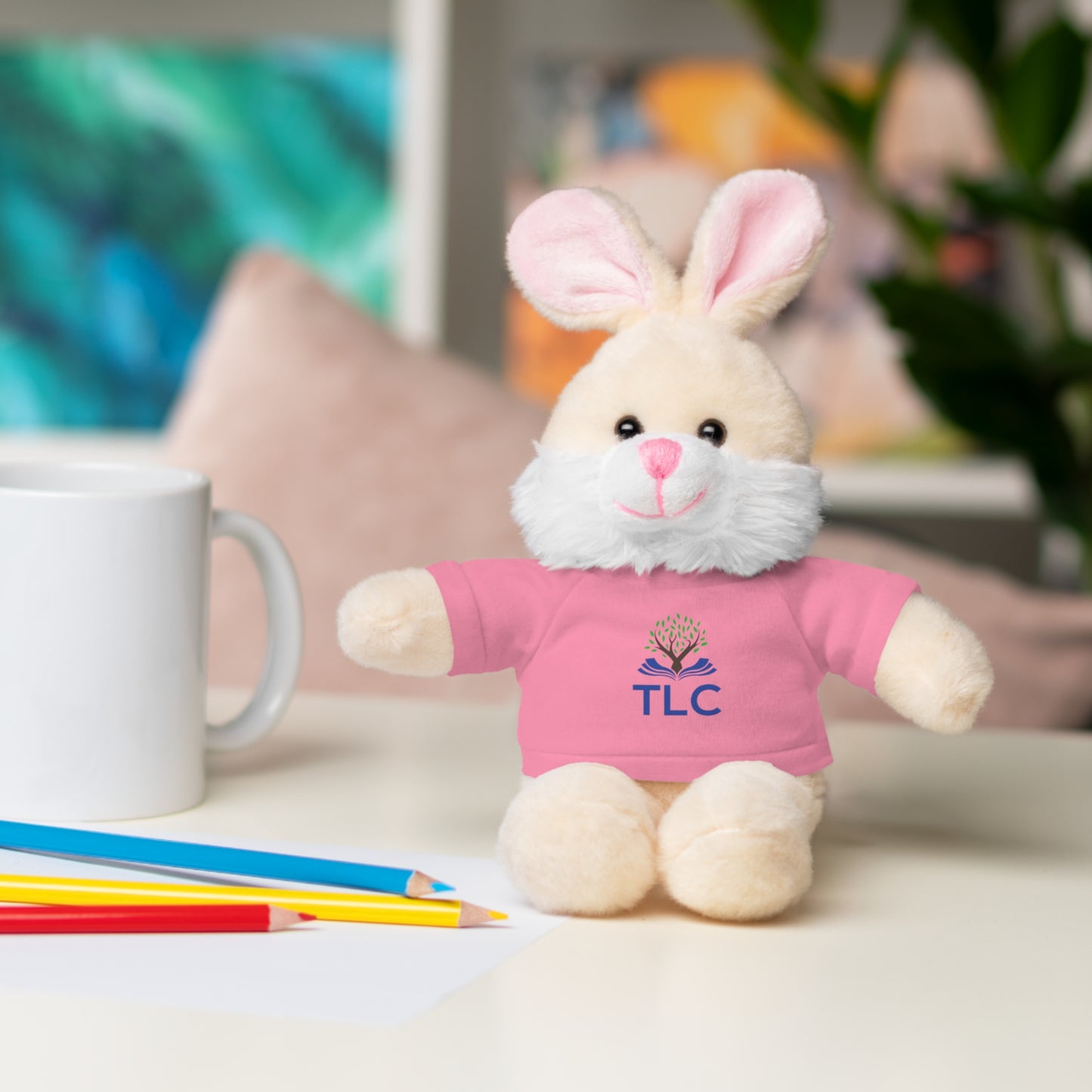 Plush bunny toy wearing a pink shirt with 'TLC' on a table with colored pencils and a mug.