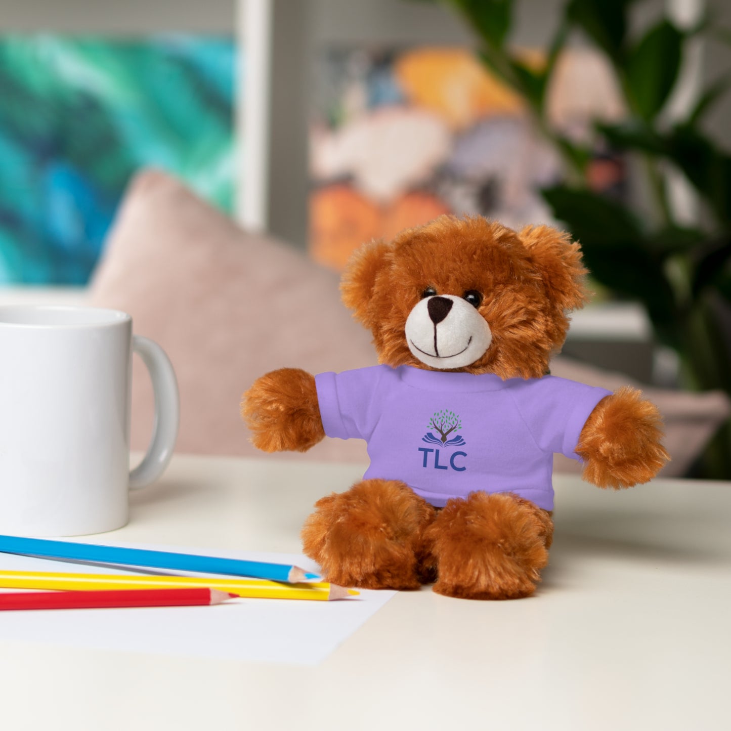 Brown teddy bear wearing a purple shirt with 'TLC' on it, sitting on a table with a mug and pencils.