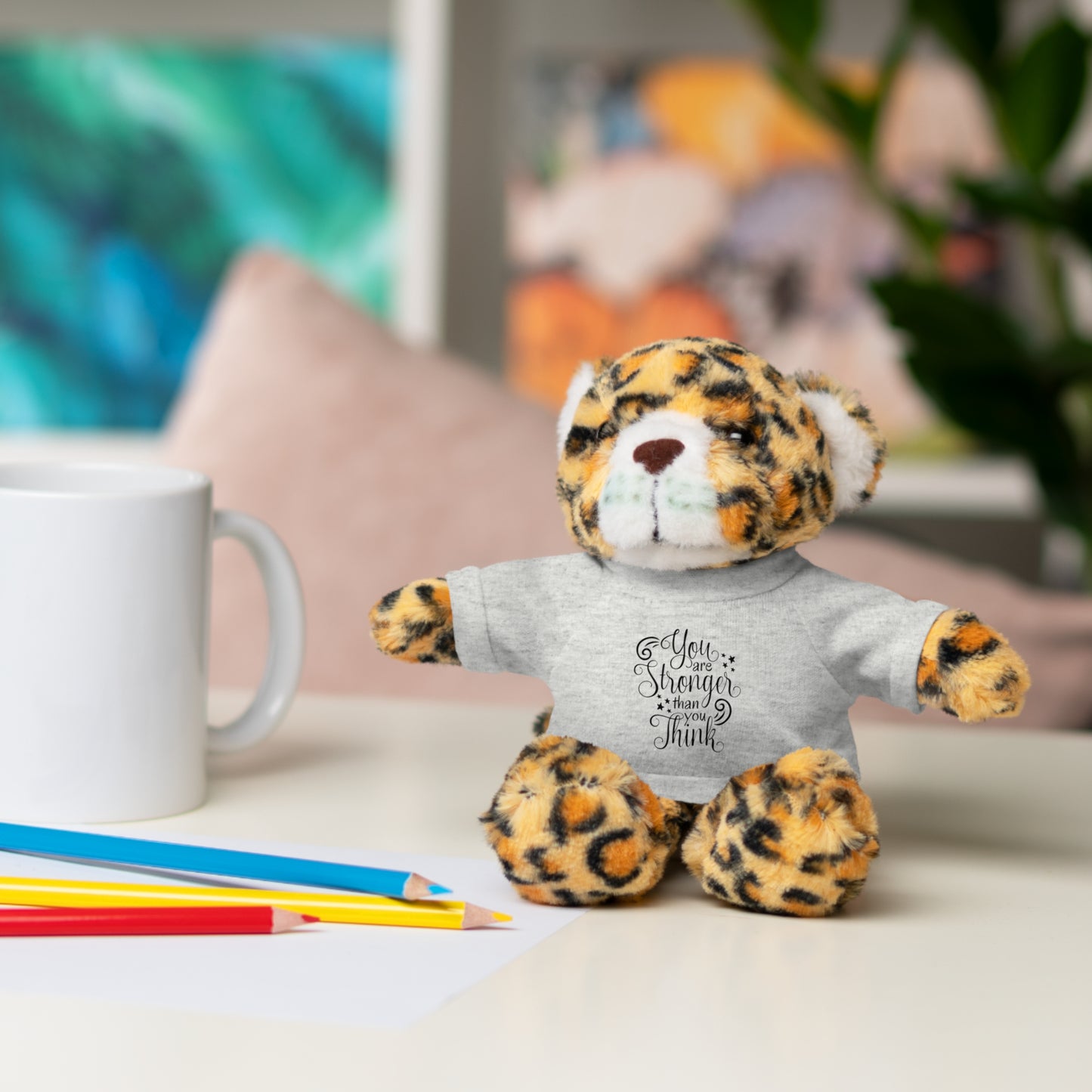 Leopard print teddy bear wearing a gray shirt with text, sitting on a desk with a mug and pencils.