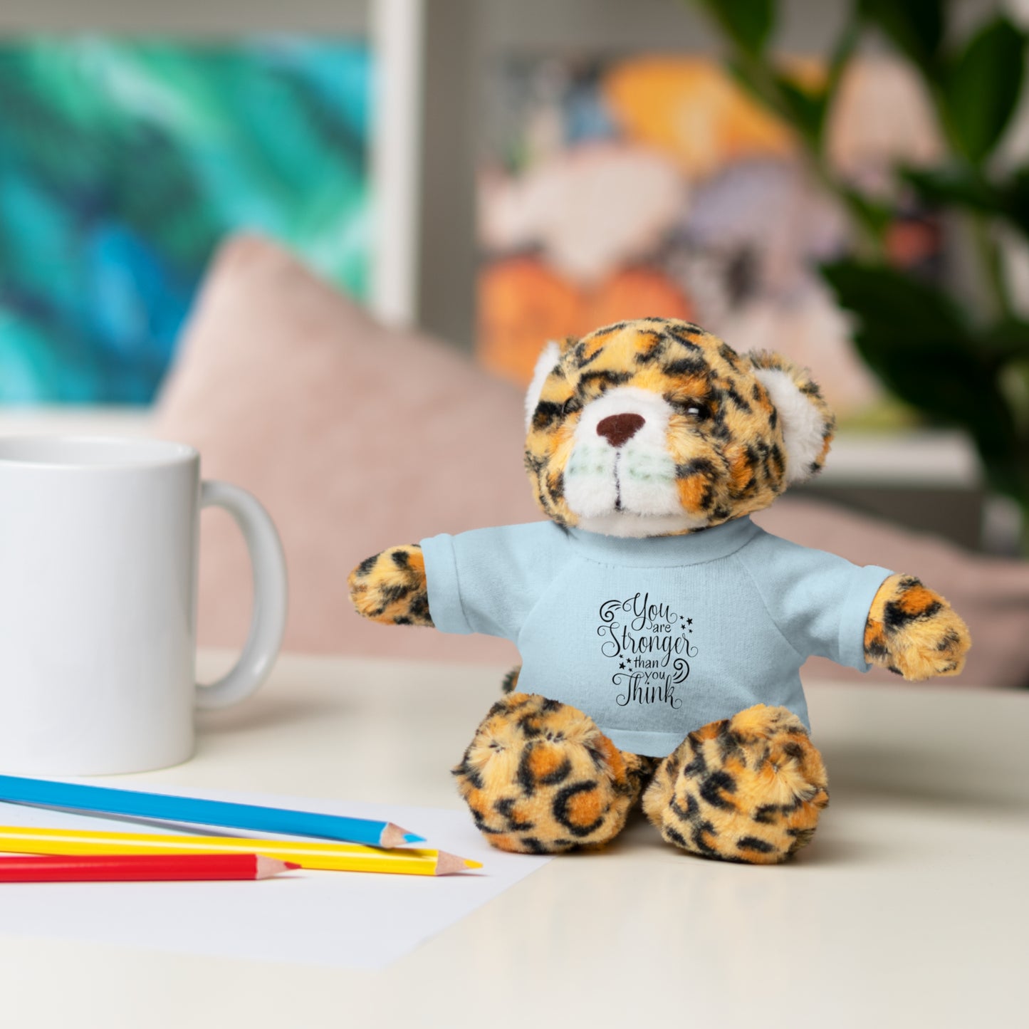 Leopard print teddy bear wearing a t-shirt with text, sitting on a table with a mug and pencils.