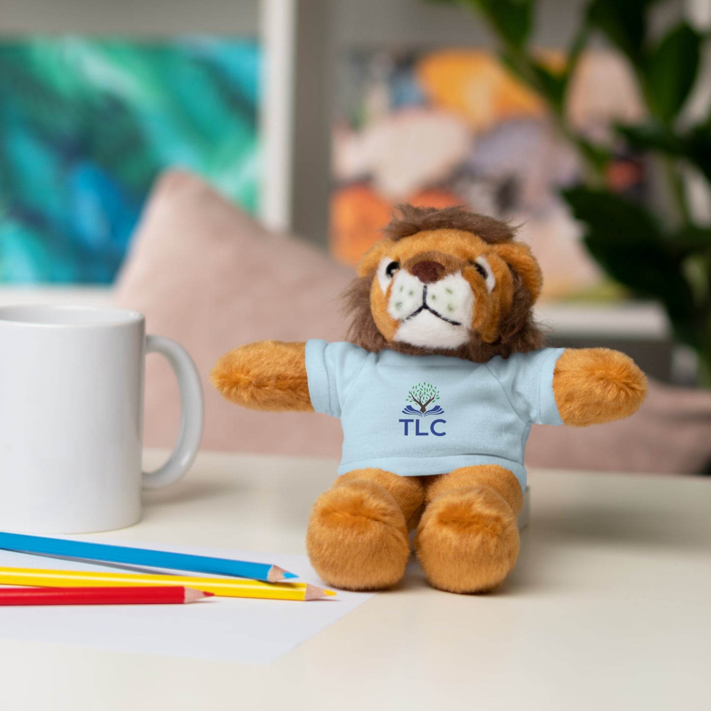 Plush lion toy wearing a blue shirt with 'TLC' logo on a table with stationery items.