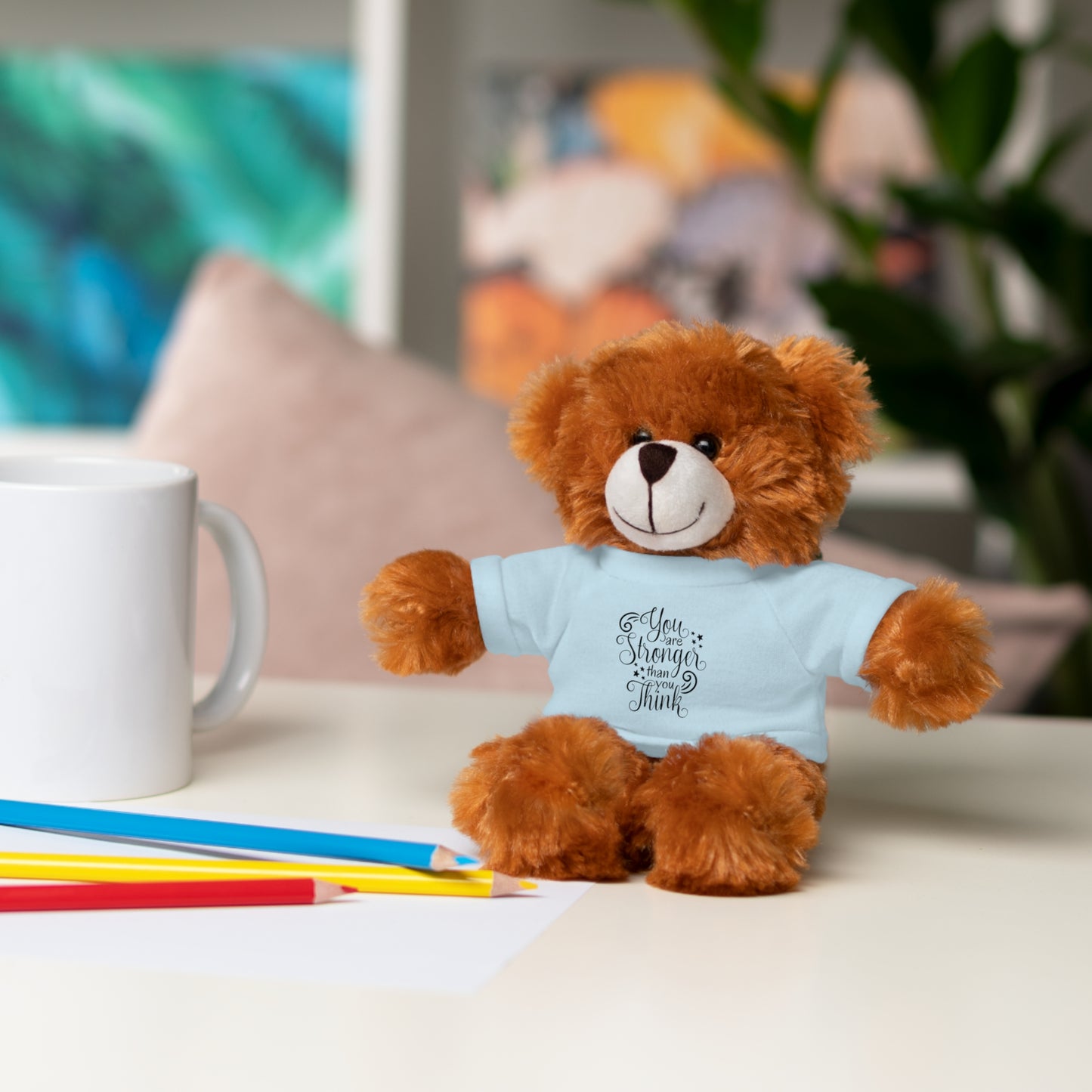 Teddy bear wearing a blue shirt with text, sitting on a table with stationery items.