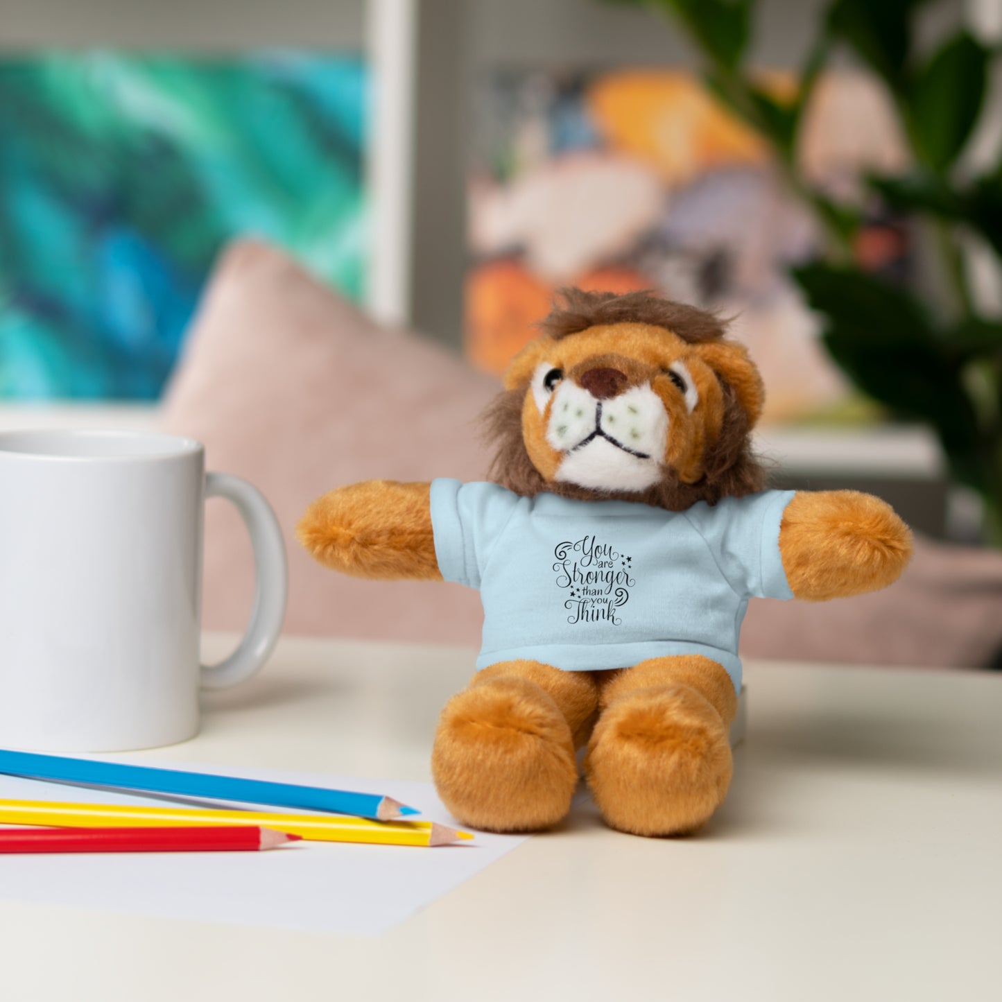Plush lion toy wearing a shirt with text, sitting on a surface with a mug and pencils in the background.