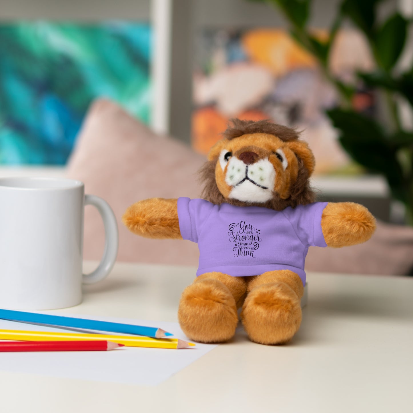 Plush lion toy wearing a purple shirt with text, sitting on a table with a mug and pencils.