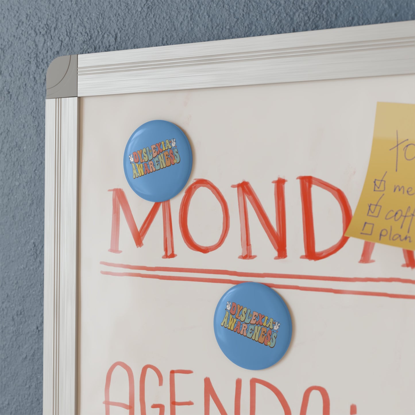 Whiteboard with 'MONDAY' and 'AGENDA' text, featuring two blue buttons with colorful text on a gray wall background.