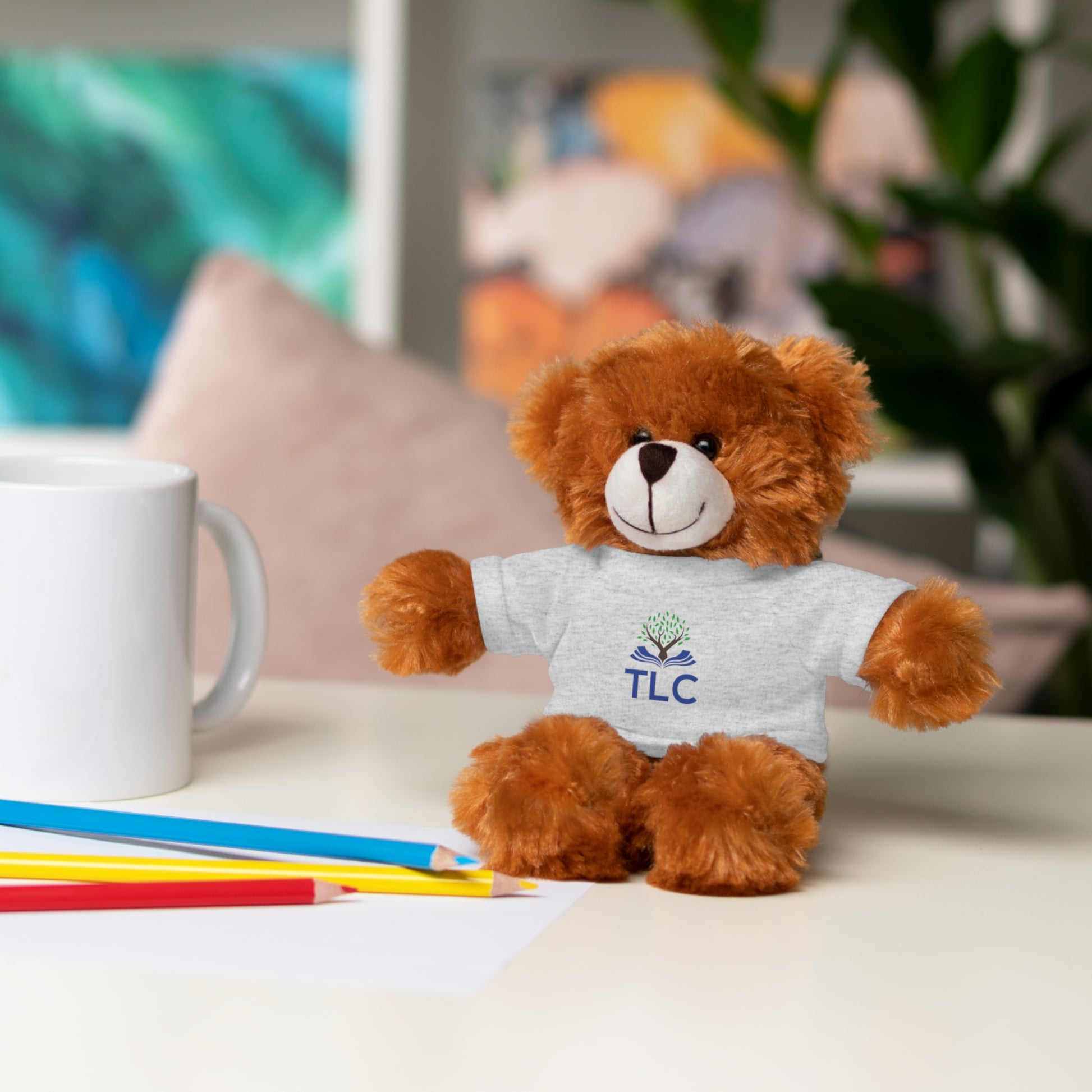 Brown teddy bear wearing a white shirt with 'TLC' logo on a table with stationery items.
