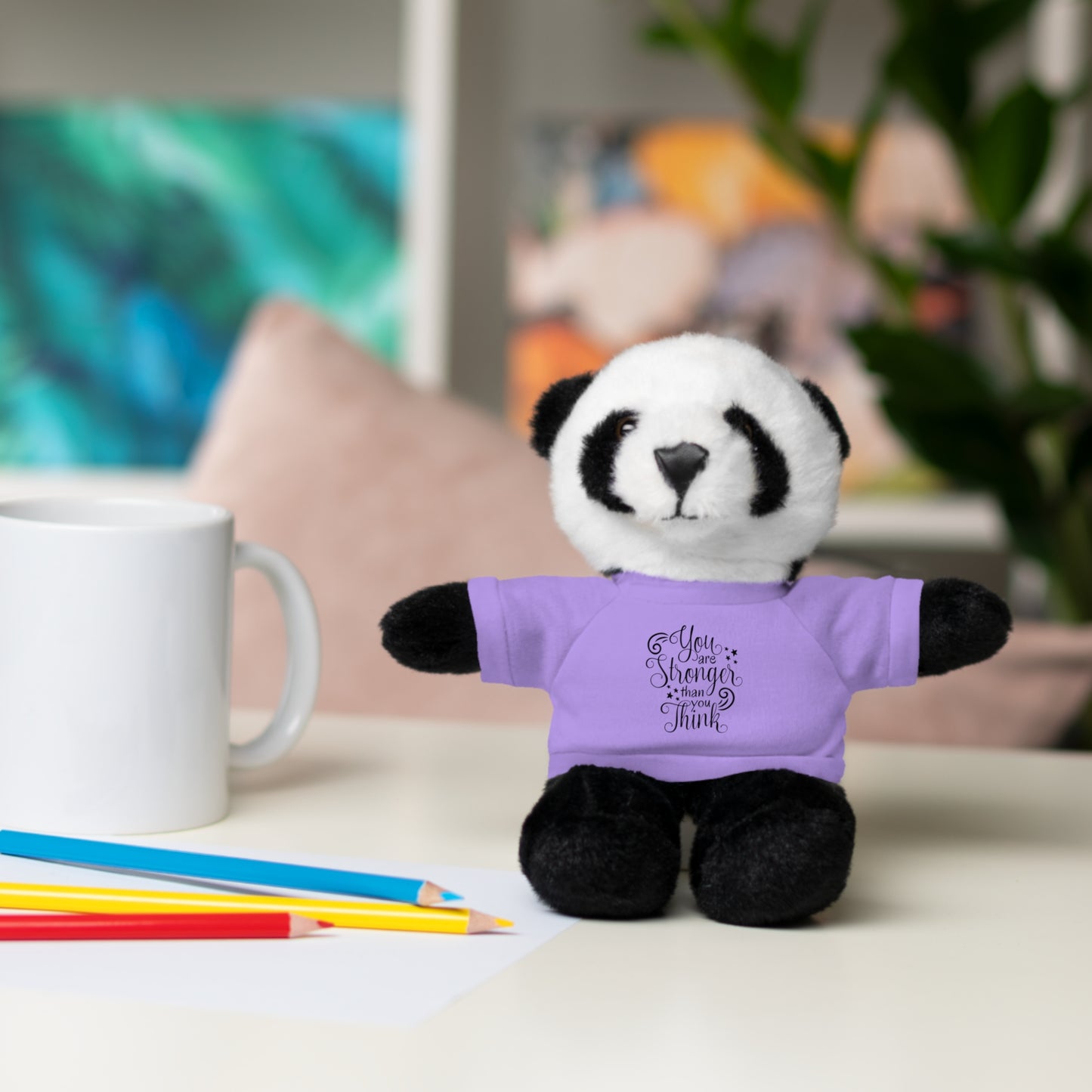 Plush panda toy wearing a purple shirt with text, placed on a surface with colored pencils and a mug.