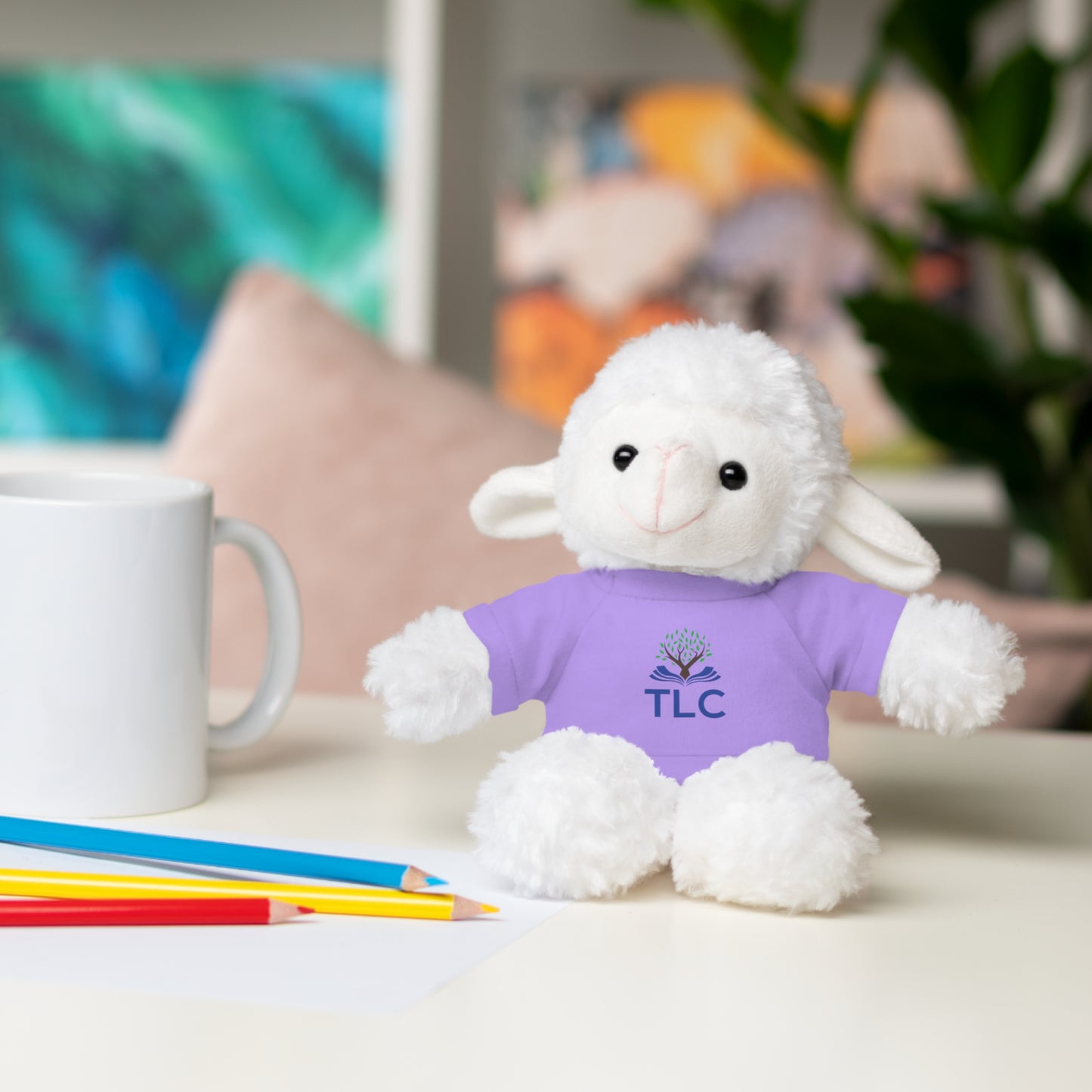White plush lamb toy wearing a purple shirt with 'TLC' logo on a table with stationery items.