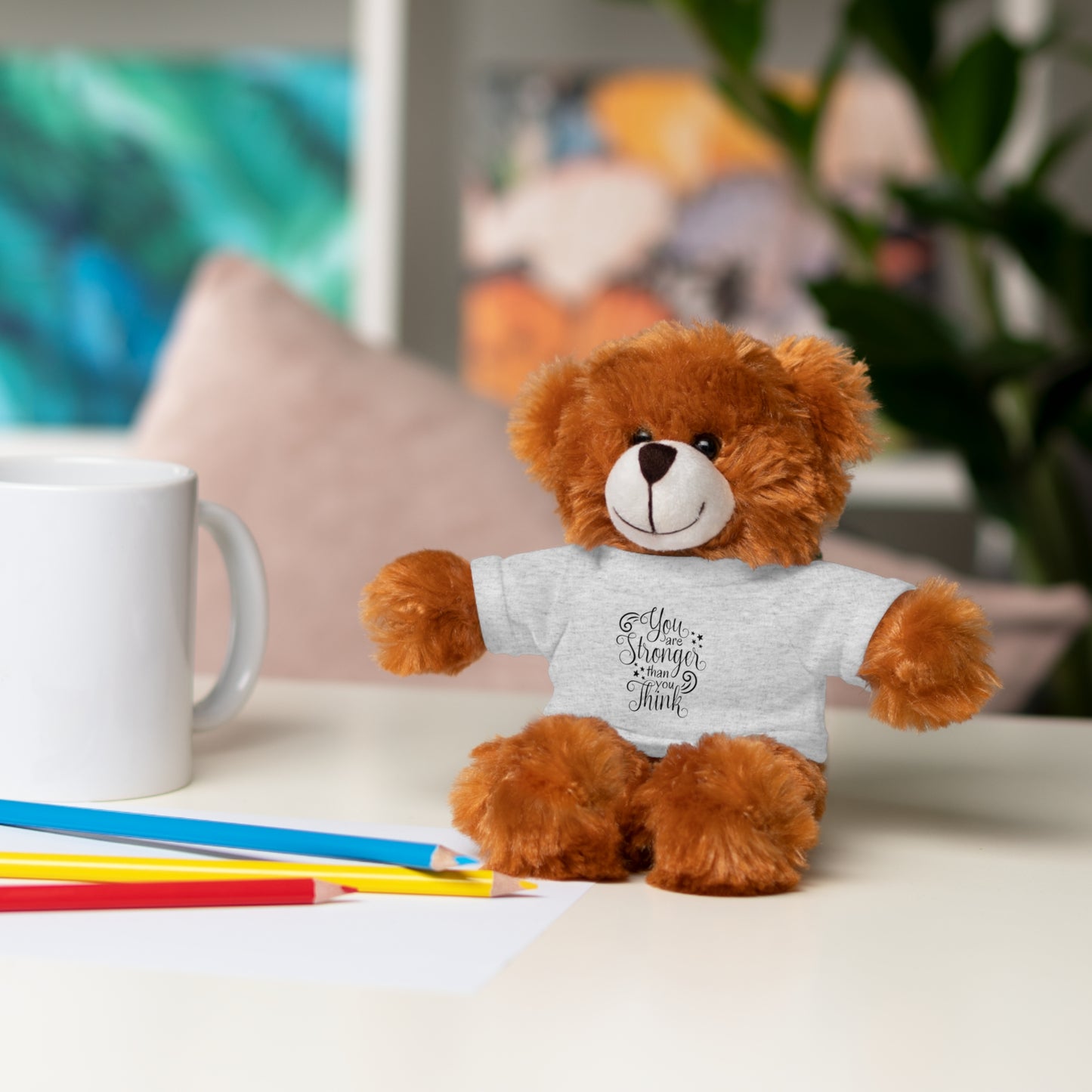 Brown teddy bear wearing a white shirt with text, sitting on a table with stationery items.