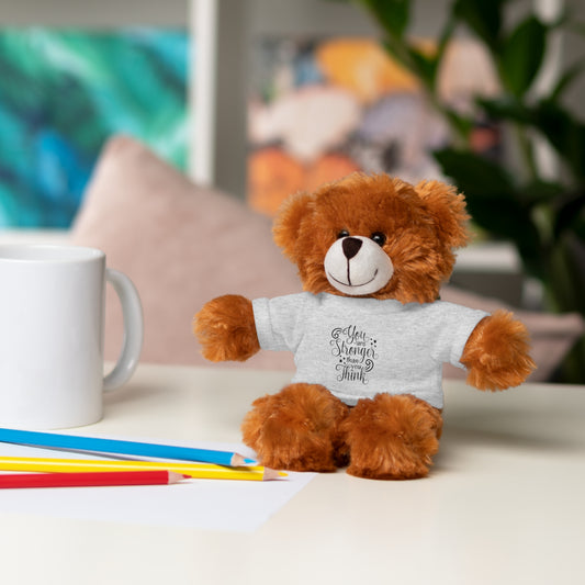 Brown teddy bear wearing a white shirt with text, sitting on a table with stationery items.
