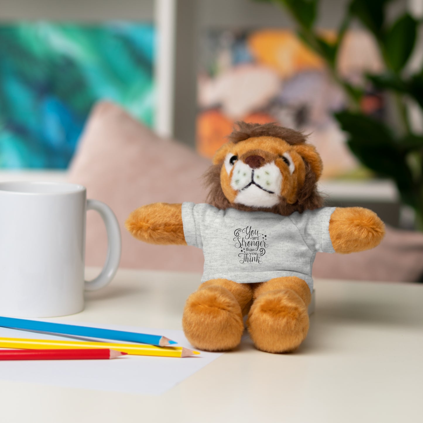 Plush lion toy wearing a shirt with text, sitting on a table with a mug and pencils.