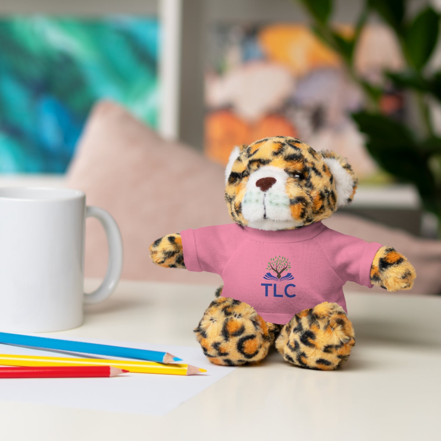 Leopard print teddy bear wearing a pink shirt with 'TLC' on it, sitting on a table with colored pencils and a mug.