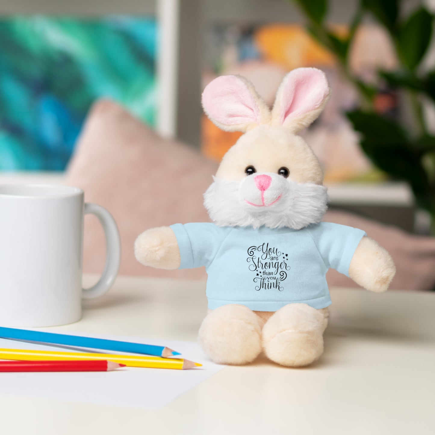 Plush bunny toy wearing a blue shirt with text, sitting on a table with a mug and pencils.