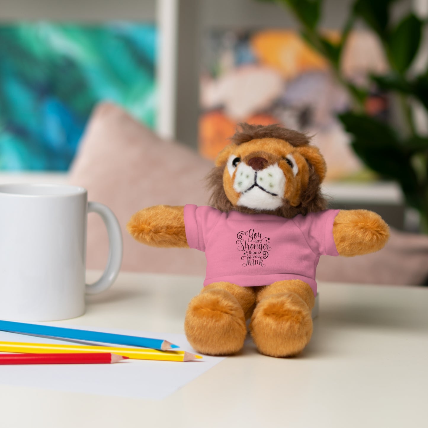Plush lion toy wearing a pink shirt with text, sitting on a surface with a mug and pencils in the background.