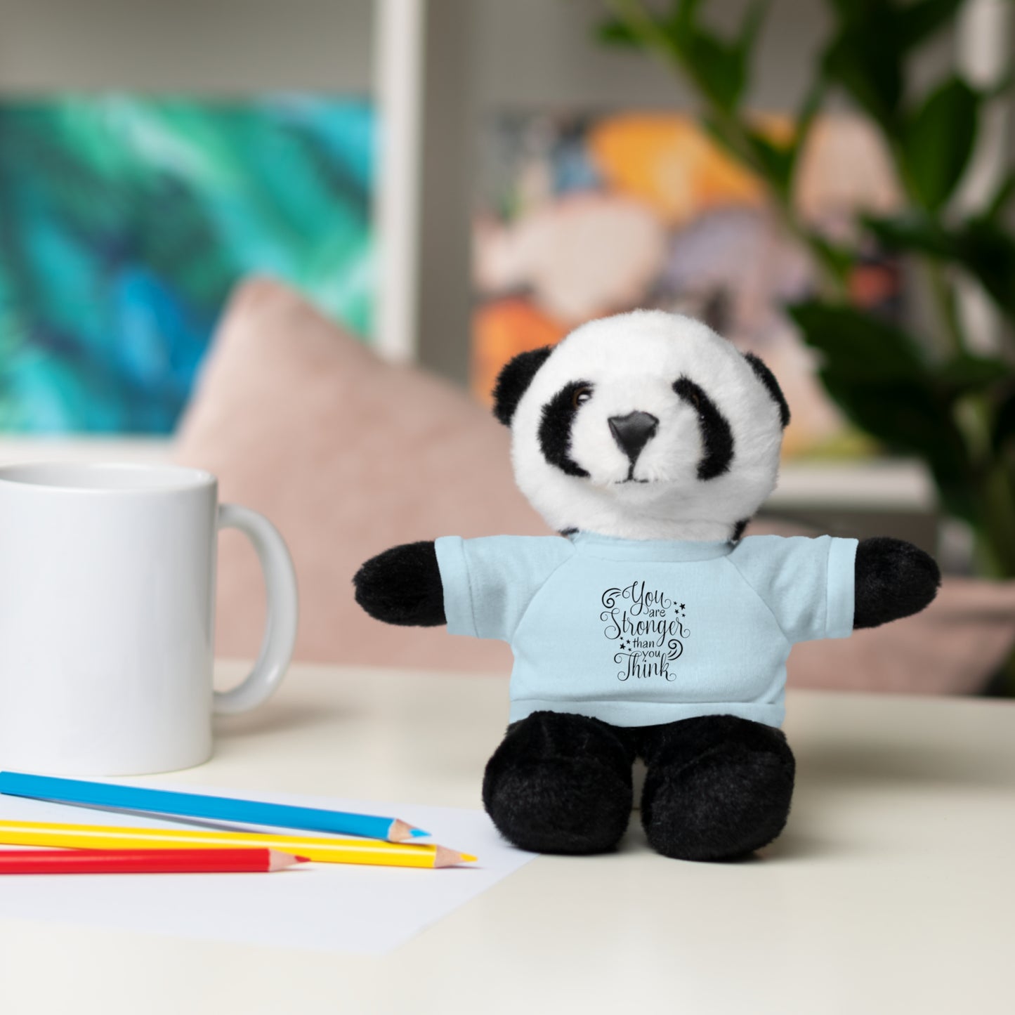 Stuffed panda toy wearing a blue shirt with text, placed on a surface with colored pencils and a mug.