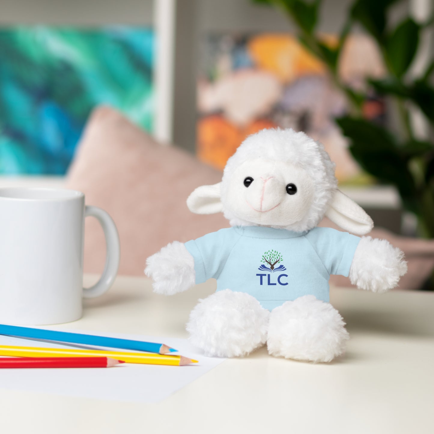 White plush lamb toy with a blue shirt featuring 'TLC' logo on a table with stationery items.