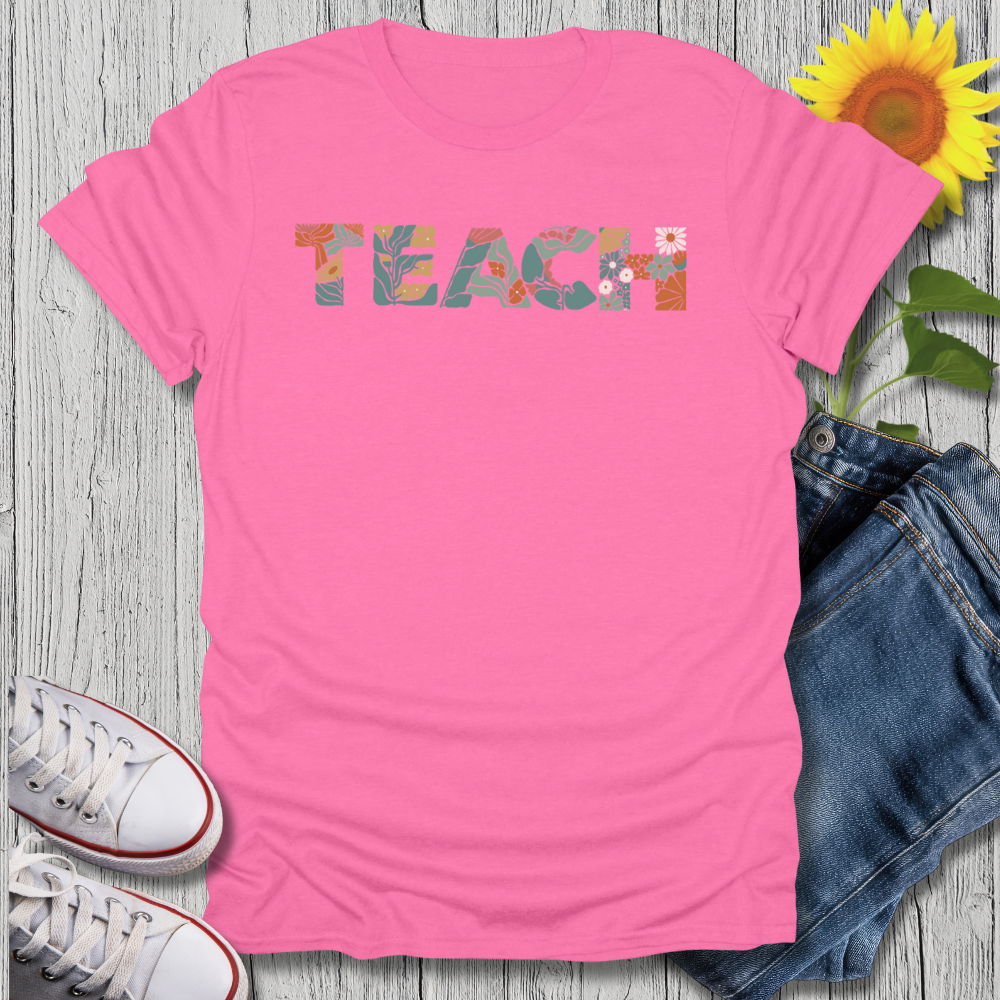 Pink t-shirt with 'TEACH' printed on it, surrounded by jeans, sneakers, and a sunflower on a wooden surface.