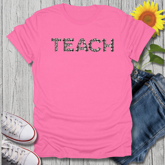 Pink t-shirt with 'TEACH' text, sunflower, jeans, and sneakers on a wooden surface