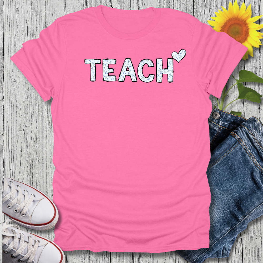 Pink t-shirt with 'TEACH' text, sunflower, jeans, and sneakers on a wooden surface