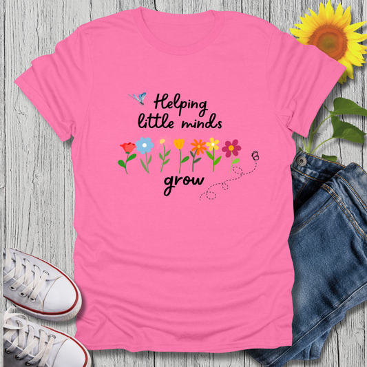 Pink t-shirt with 'Helping little minds grow' text and flowers on a wooden surface with jeans and sneakers.