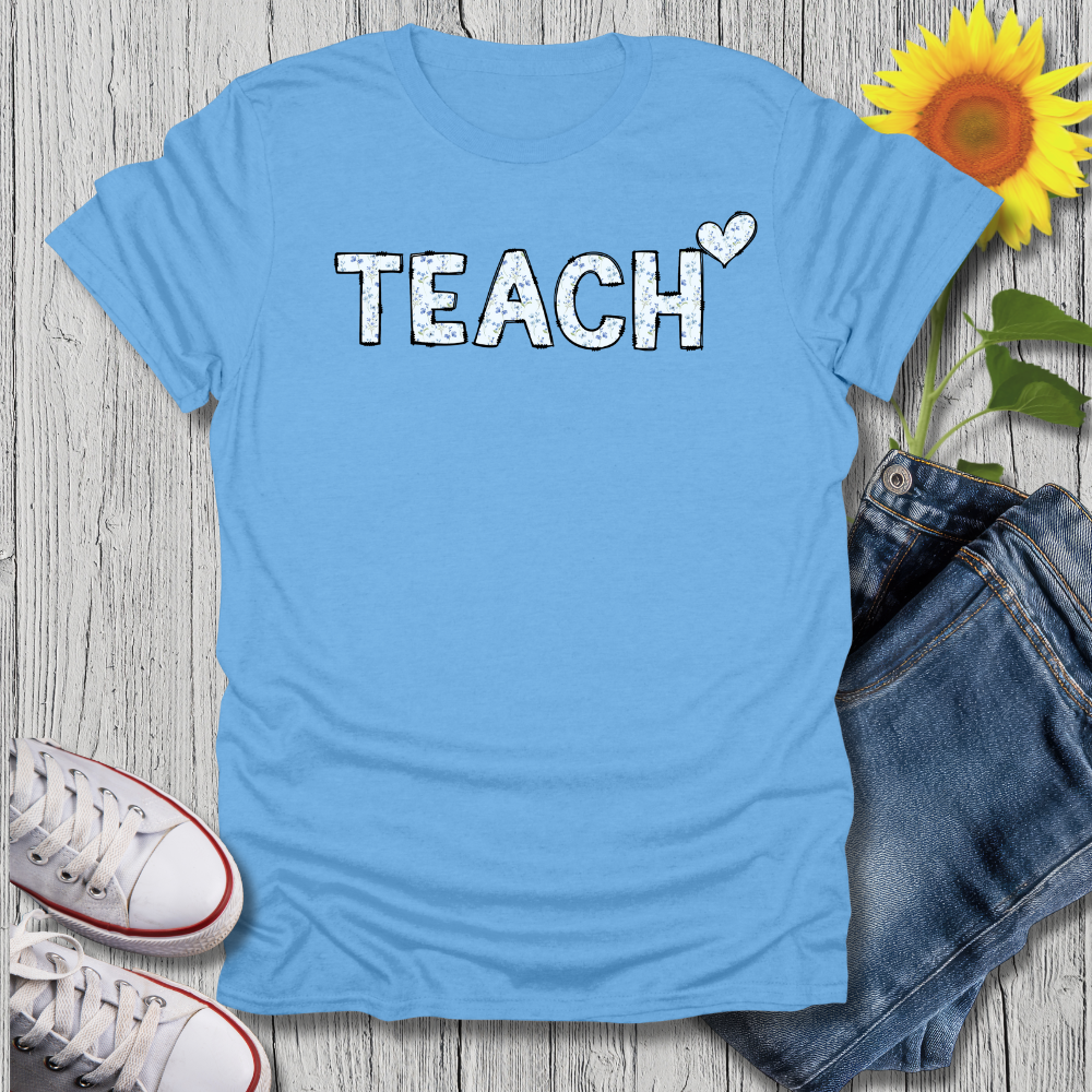 Blue t-shirt with 'TEACH' text, sunflower, jeans, and sneakers on a wooden surface