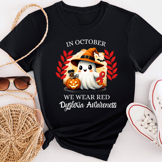 Black T-shirt featuring a cute Halloween ghost wearing a witch hat, holding a red dyslexia awareness ribbon and a coffee cup. The ghost is surrounded by red autumn leaves, a carved pumpkin, and bats. White text reads: “In October We Wear Red Dyslexia Awareness.