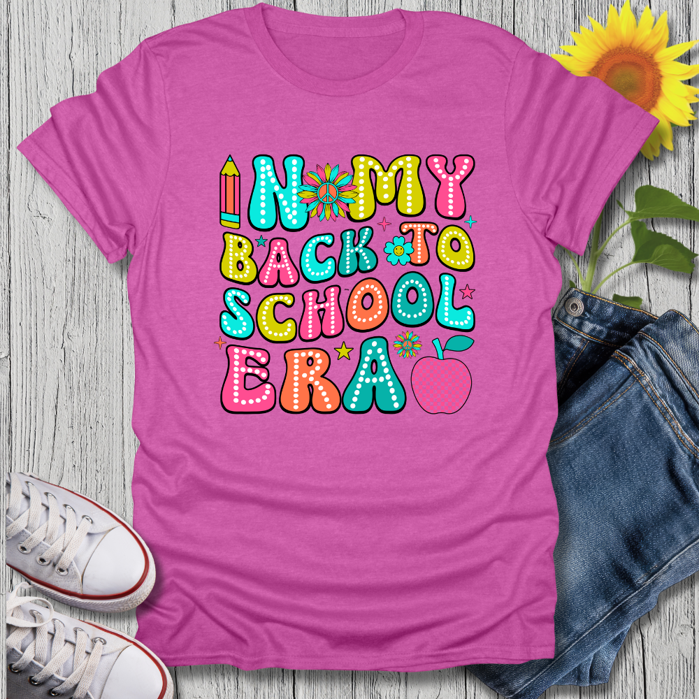 Pink t-shirt with colorful 'Back to School' text, sunflower, jeans, and sneakers on a wooden surface