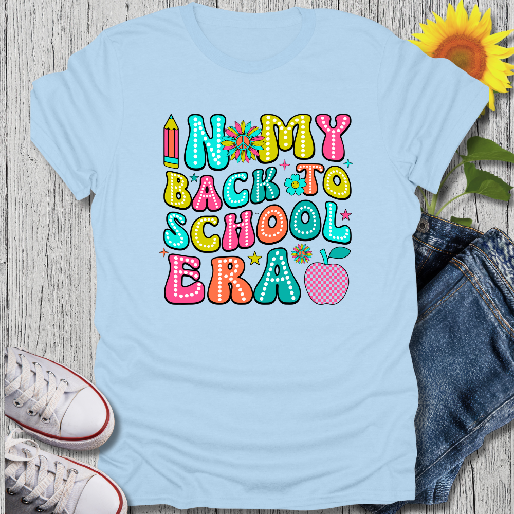 Light blue t-shirt with colorful 'Back to School' text design on a wooden surface with jeans, sneakers, and a sunflower.