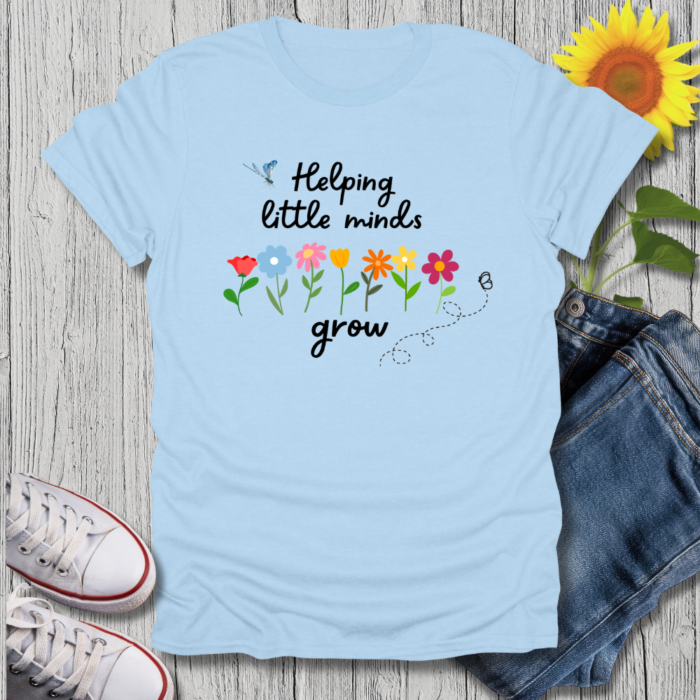 Light blue t-shirt with 'Helping little minds grow' text and flowers on a wooden surface with jeans and sneakers.
