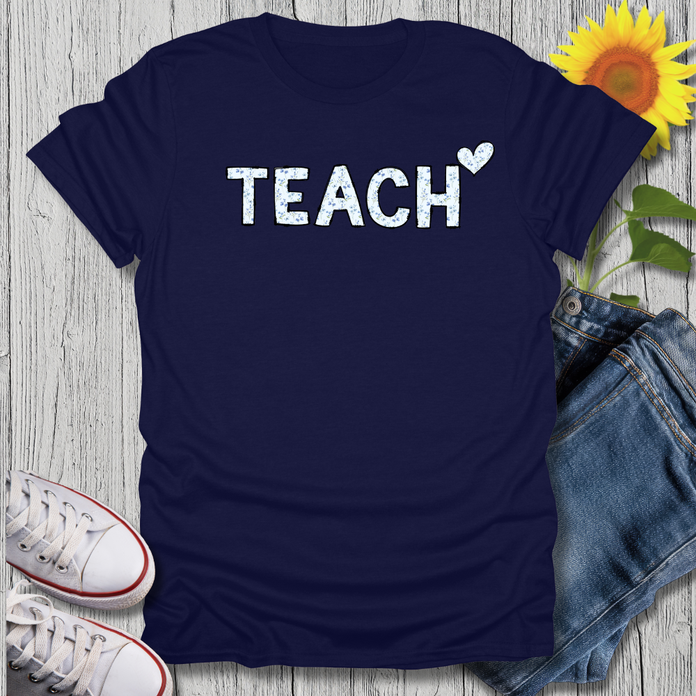 Navy blue t-shirt with 'TEACH' print on a wooden surface with jeans and sneakers.