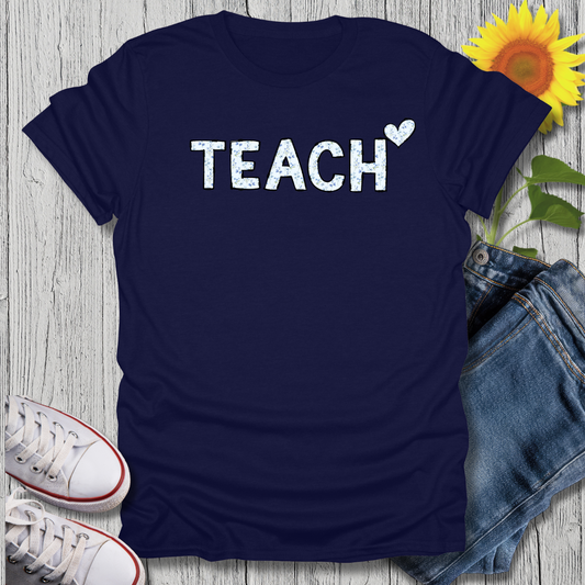 Navy blue t-shirt with 'TEACH' print on a wooden surface with jeans and sneakers.
