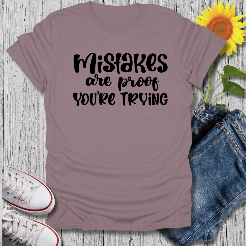 Mauve t-shirt with 'Mistakes are proof you're trying' text, paired with jeans and sneakers on a wooden surface.