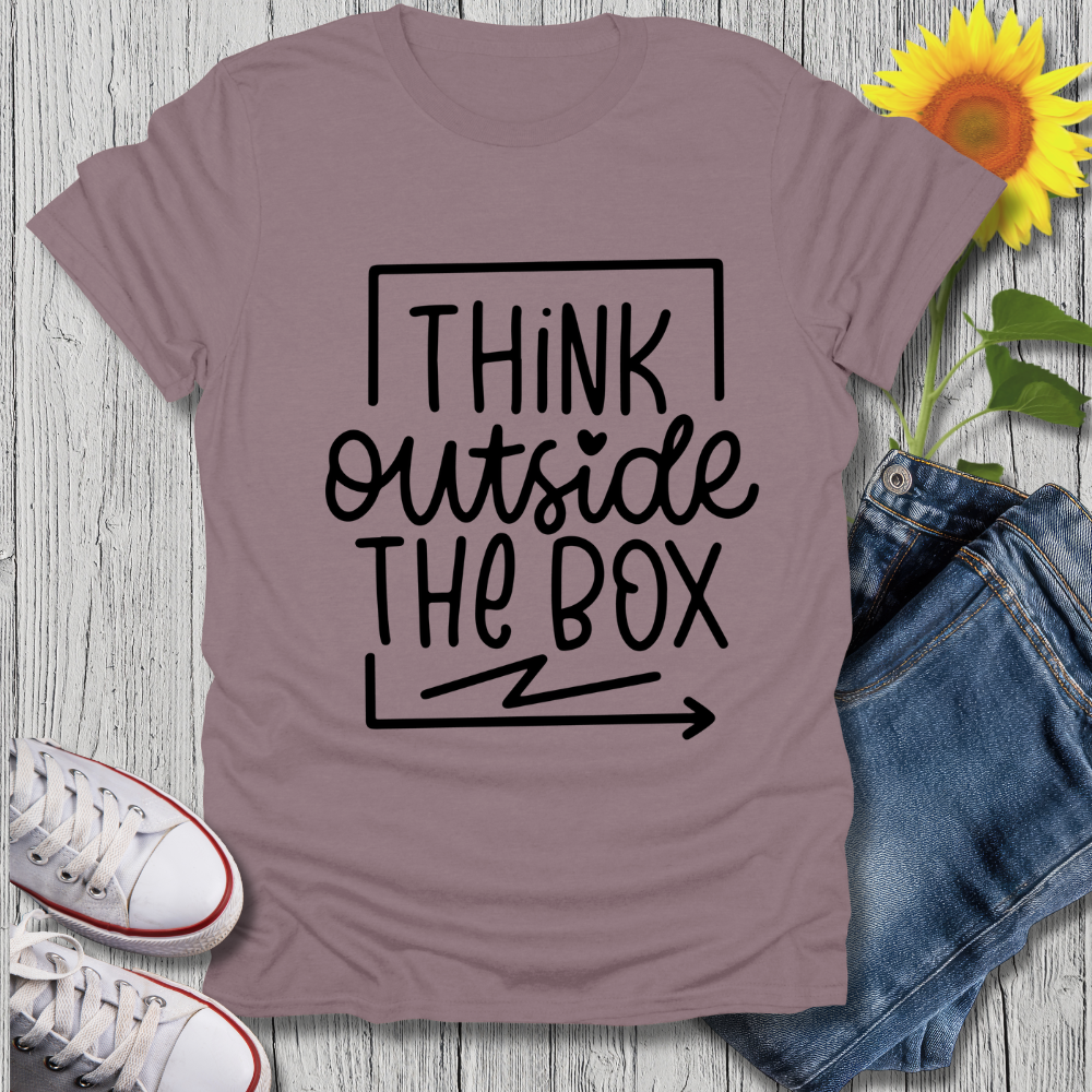Mauve t-shirt with 'Think Outside The Box' text, paired with jeans and sneakers on a wooden surface.