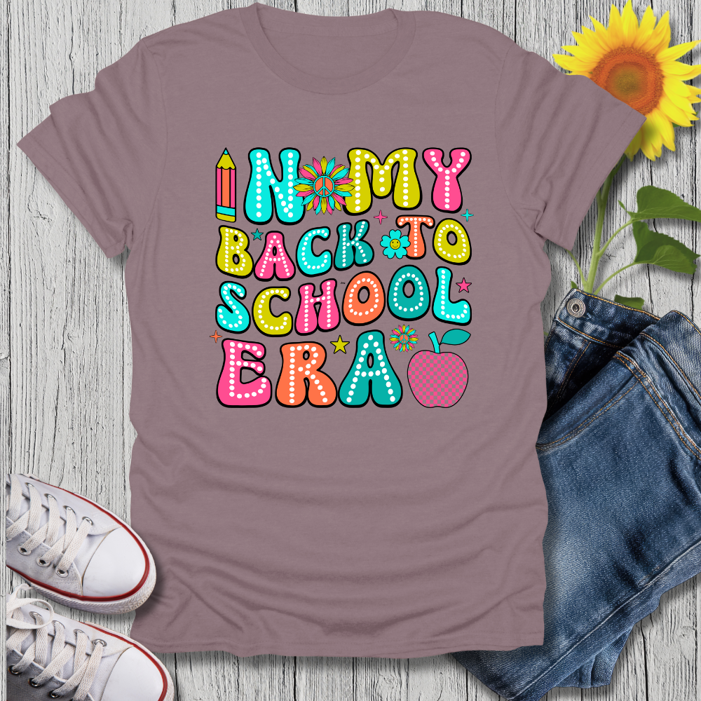 T-shirt with 'In My Back to School Era' text on a wooden surface with jeans and sneakers.