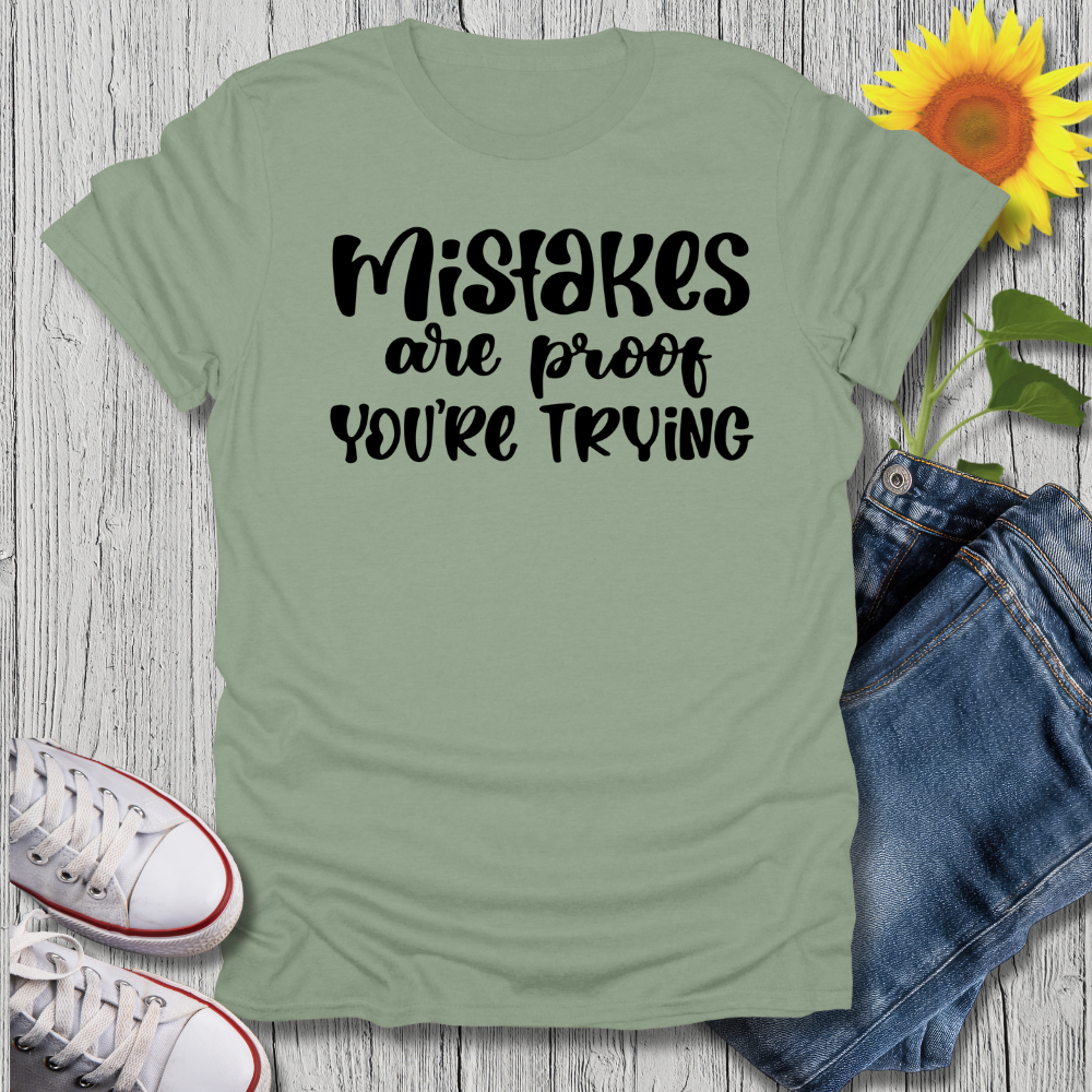 Green t-shirt with 'Mistakes are proof you're trying' text, paired with jeans and sneakers on a wooden surface.