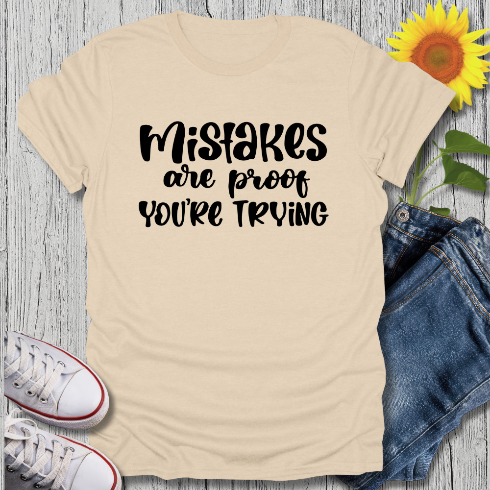 Beige t-shirt with 'Mistakes are proof you're trying' text, paired with jeans and sneakers on a wooden surface.