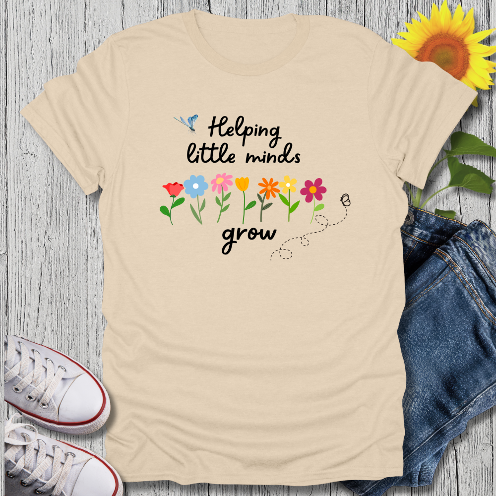 Beige t-shirt with 'Helping little minds grow' text and flowers on a wooden surface with jeans and sneakers.