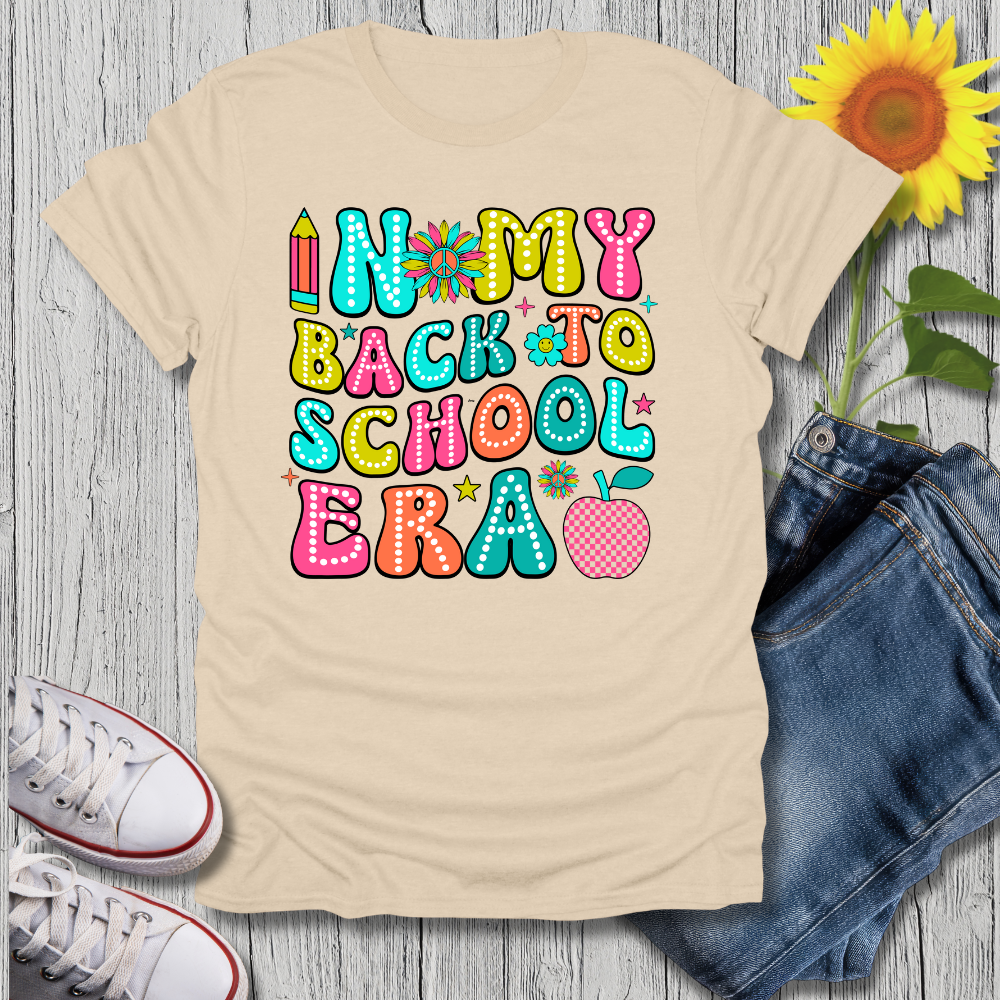 Beige t-shirt with colorful 'In My Back to School Era' text, white sneakers, blue jeans, and a sunflower on a wooden surface.
