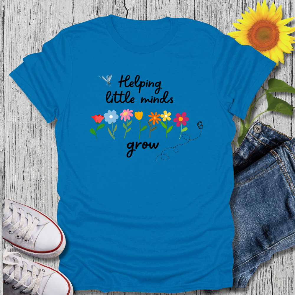 Blue t-shirt with 'Helping little minds grow' text and flowers on a wooden surface with sneakers and jeans.
