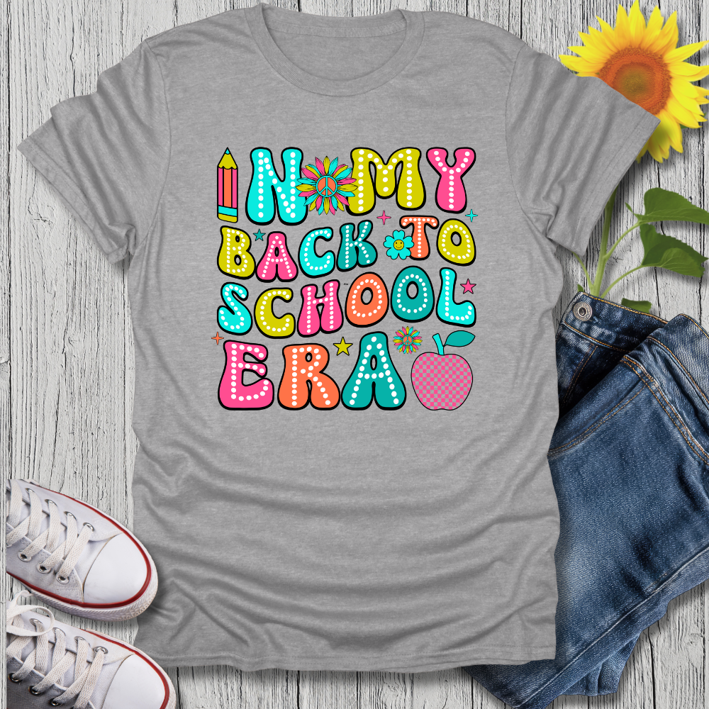 Gray t-shirt with colorful 'Back to School' text design on a wooden surface with jeans, sneakers, and a sunflower.