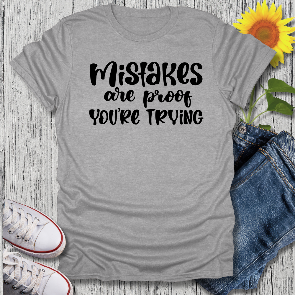 Gray t-shirt with 'Mistakes are proof you're trying' text, paired with jeans and sneakers on a wooden surface.