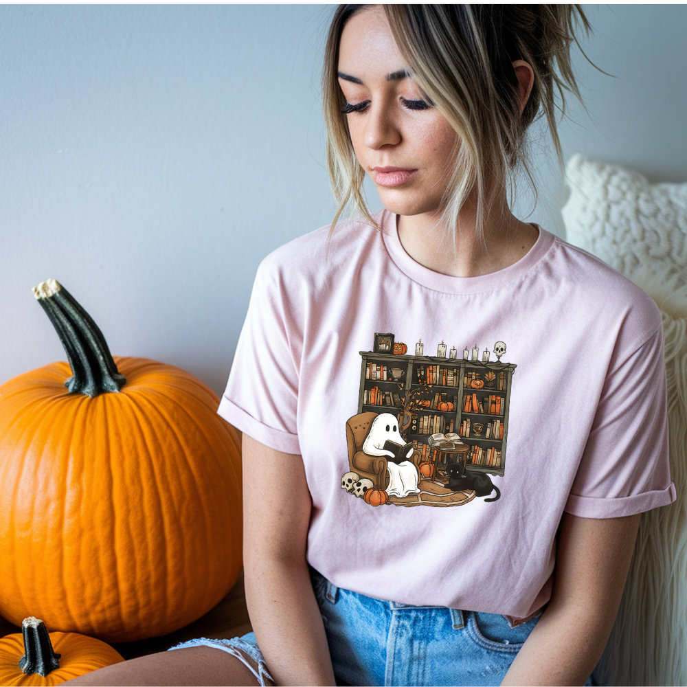 Woman wearing a pink t-shirt with a ghost and bookshelf design, sitting next to pumpkins.