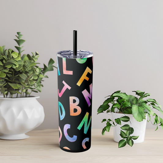 Tumbler with colorful letters on a black background, placed on a surface with plants in the background.